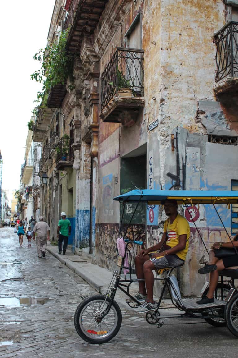 at a corner of an old street in Havana. The walls of the buildings are slightly damaged. There is a man sitting on this bike on the corner