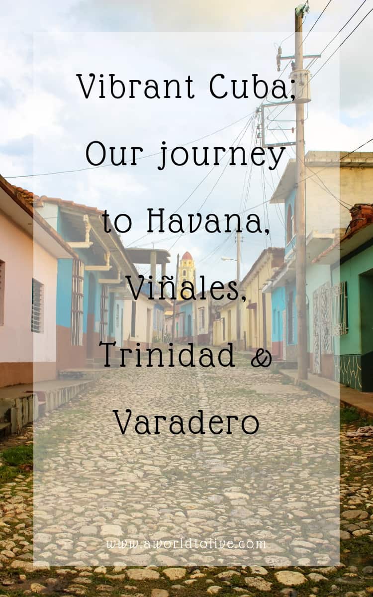 A narrow cobble stone street, lined with one level colorful buildings. A long yellow bell tower is at the end of the street. The image is covered in text saying; Visiting Cuba & Exploring Havana, Viñales, Trinidad & Varadero