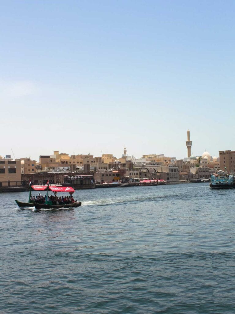 An image of a river with old buildings in the distance, a wooden boat sails by. In this Dubai travel guide there is information on shopping in this area.