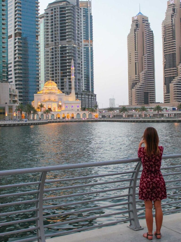 Elyse standing on the waters edge looking across to the other side, tall skyscrapers and a large mosque. The Dubai mosque has bright lights turned on.