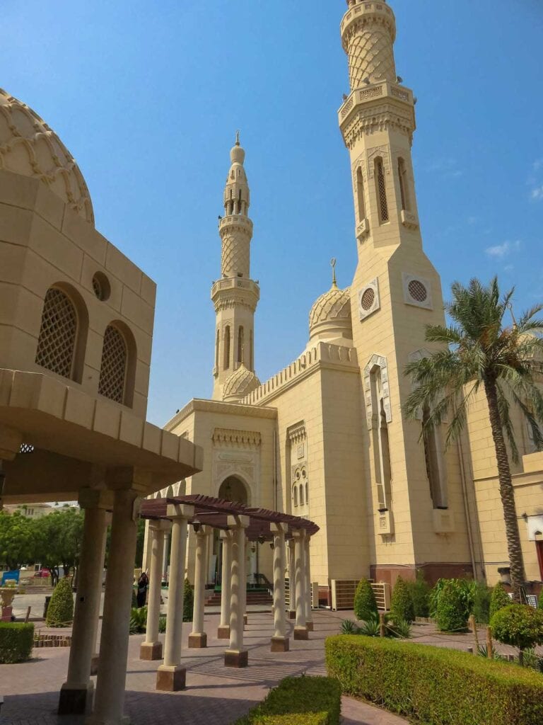 On a sunny day and image of Jumeirah Mosque. The Mosque is a yellow color and surrounded by green plants. I recommend visiting this mosque as mentioned in this Dubai Travel Guide.