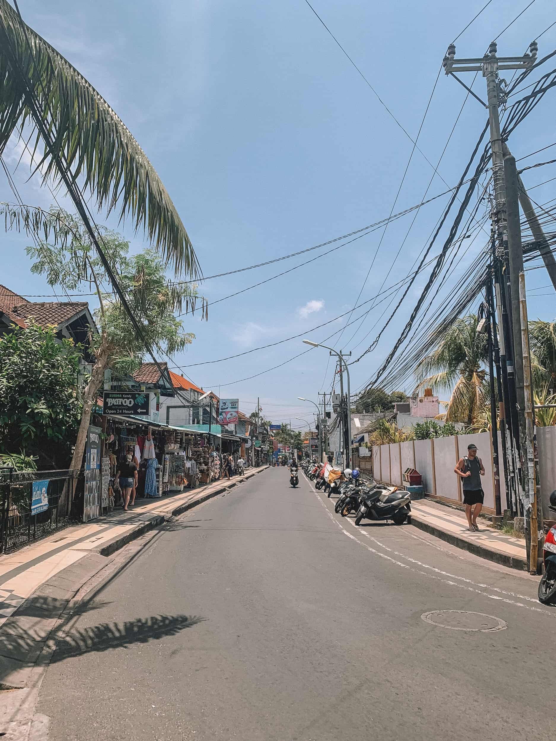 Taken down a street in Bali. The road is empty except 1 bike. There are scooters parked on the right side and local shops to the left