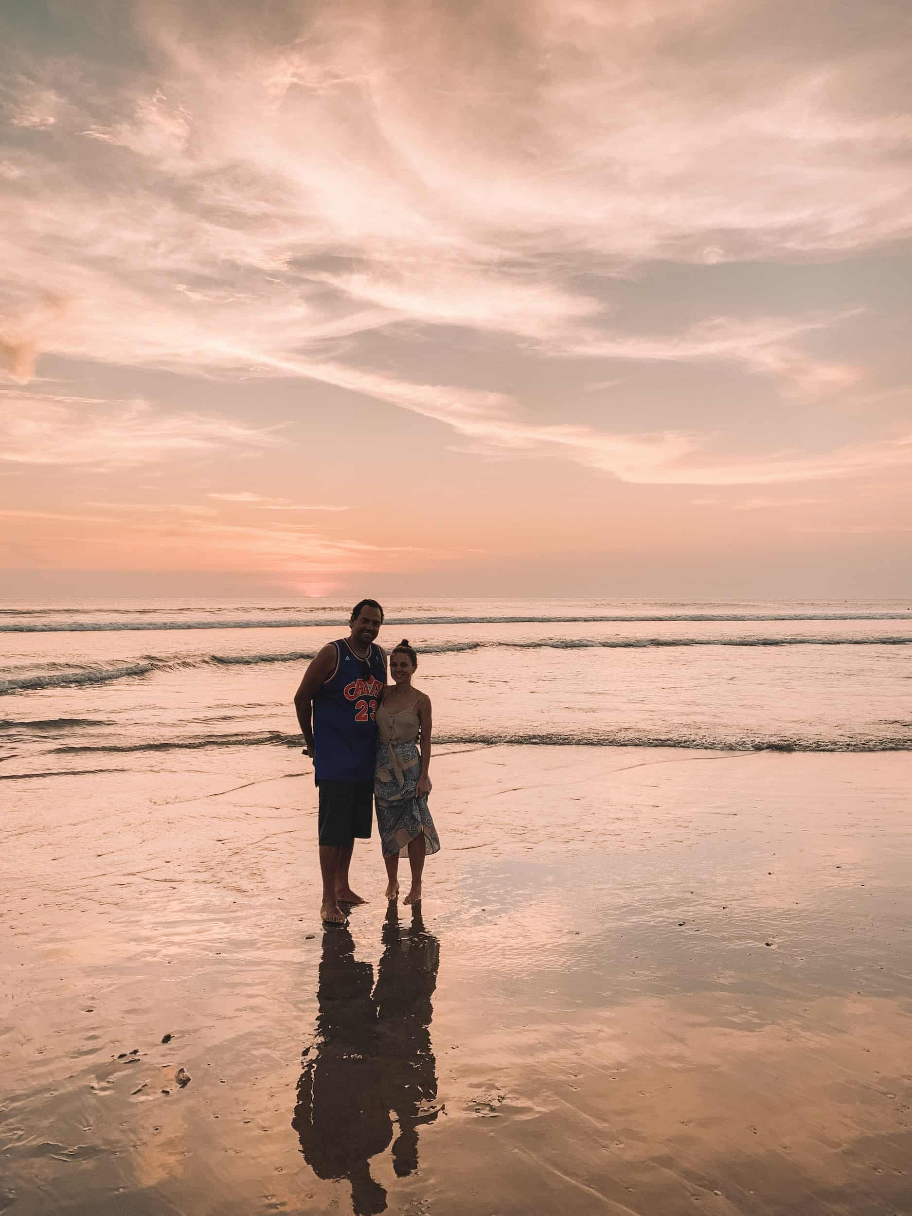A peach colored sunset shines behind a man and a woman on the beach in Bali