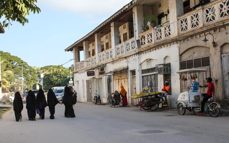 An old, white, two story building to the left. Two local men are selling drinks and bananas. Five women are walking in a row