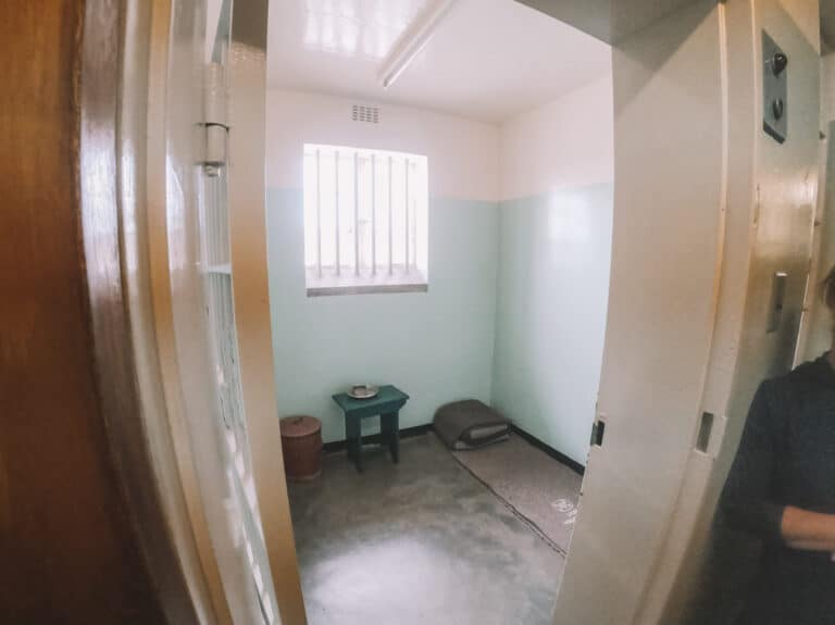 From the doorway of a prison cell. The cell only has a blanket on the floor, a side table and a metal cup and plate.