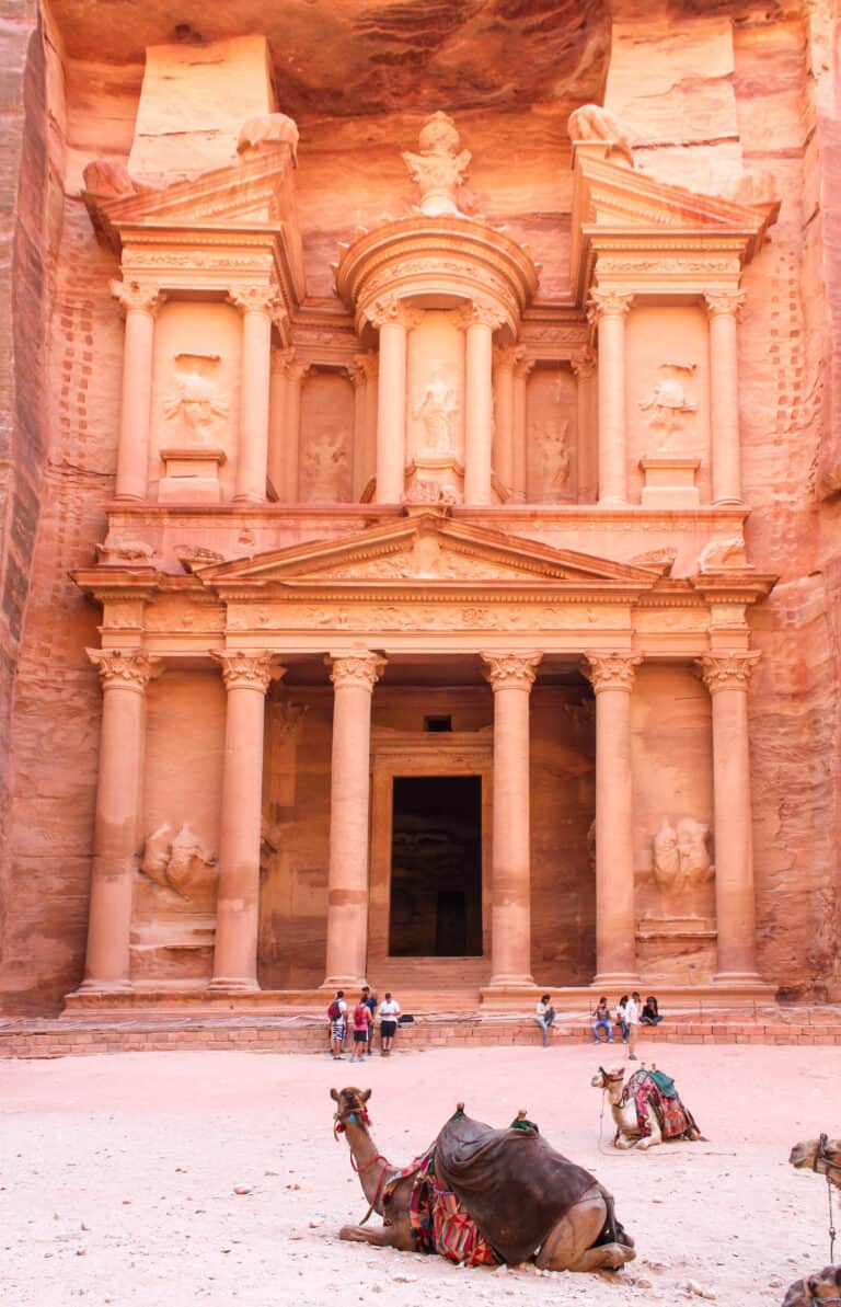 A large building carved out of orange stone. Three camels sit on the ground in front