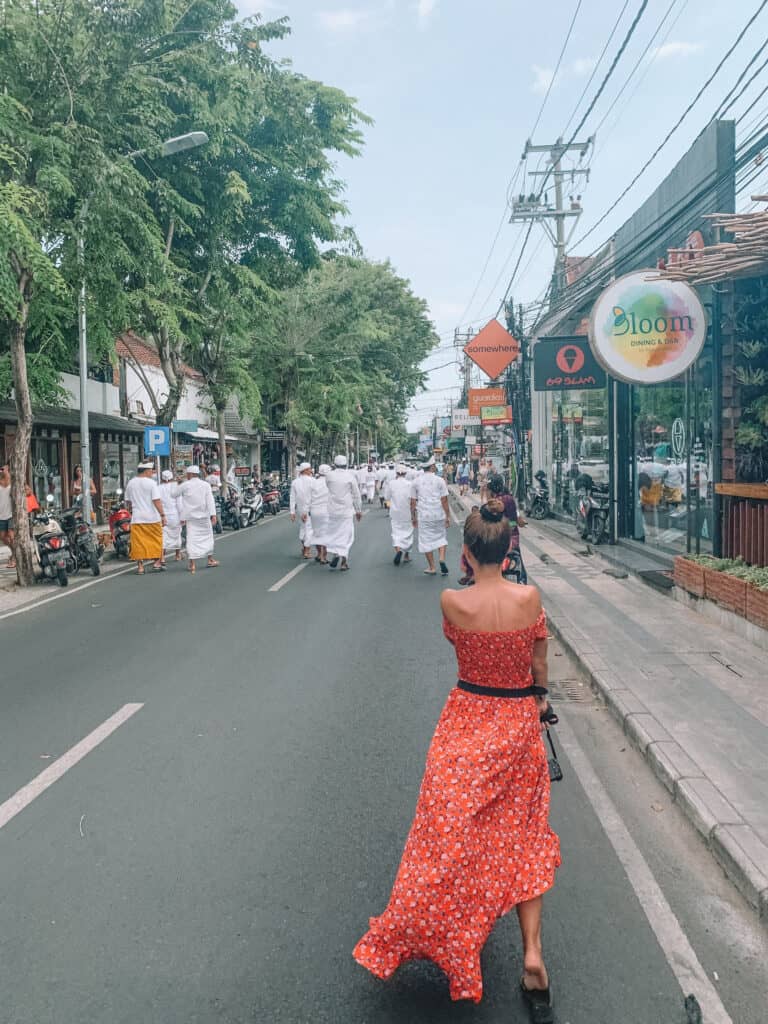 elyse walks down the street in Seminyak, wearing a bright red dress, Ahead of her is local people wearing white