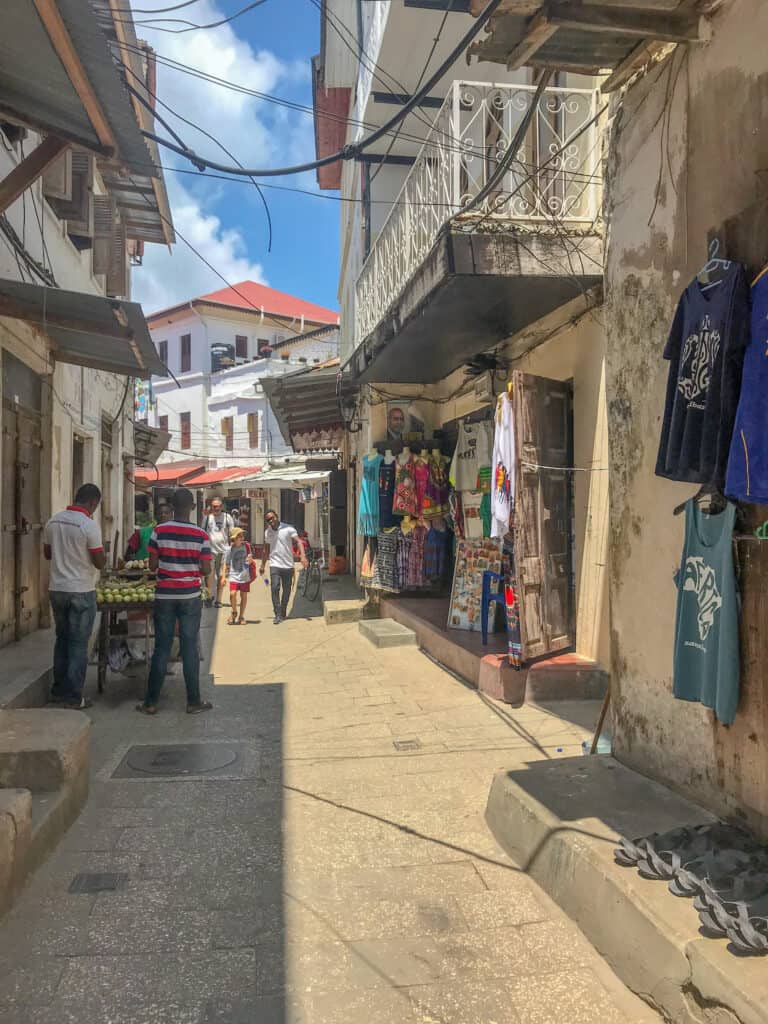 Stone Town street filled with local stores selling clothes and fruit, in the distance there's a few people