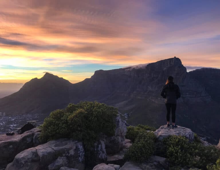 Taken at sunrise the sky is yellow, purple and blue. One female stands on the cliffs edge looking towards Table Mountain in cape town