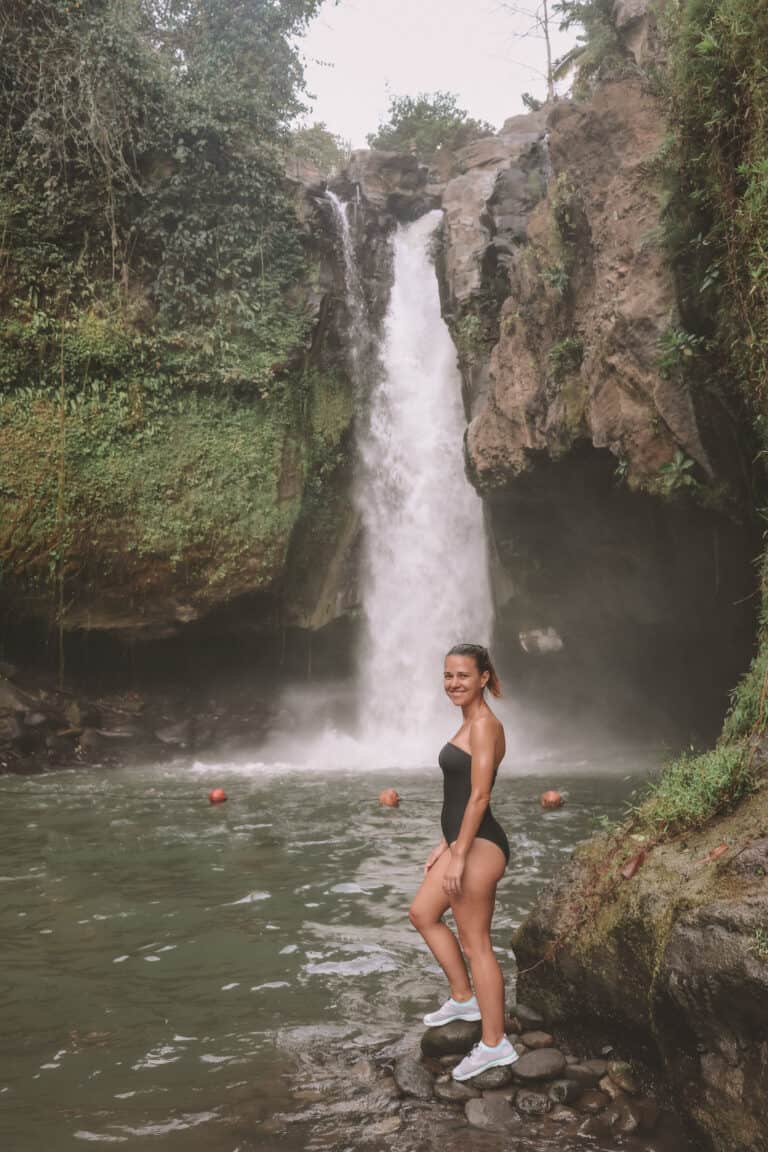 elyse standing on wet rocks at Tegenungan Village Waterfall during travel in Bali. There is a heavy flowing waterfall in the background