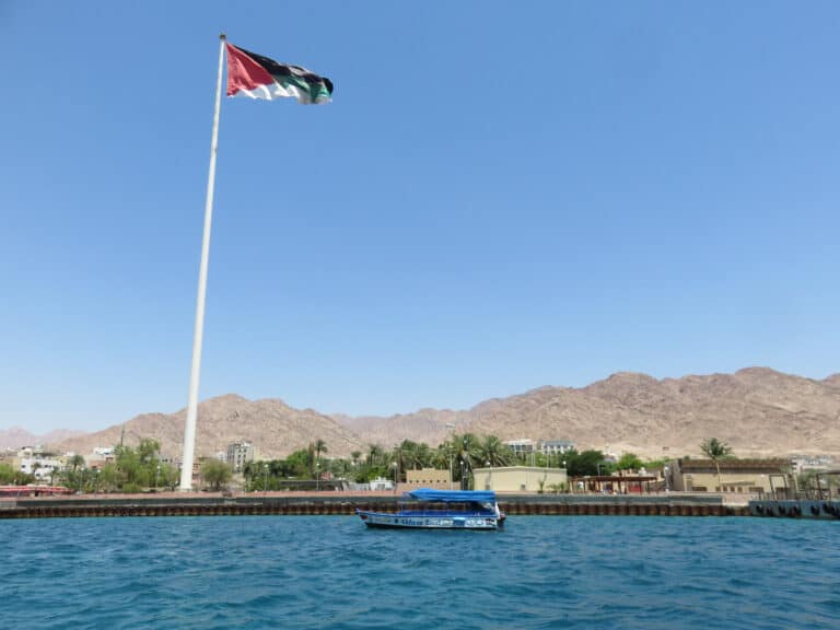 Taken on the Red Sea, land is in the distance with palm trees and mountains. A large flag pole is to the left with Jordans national flag attached