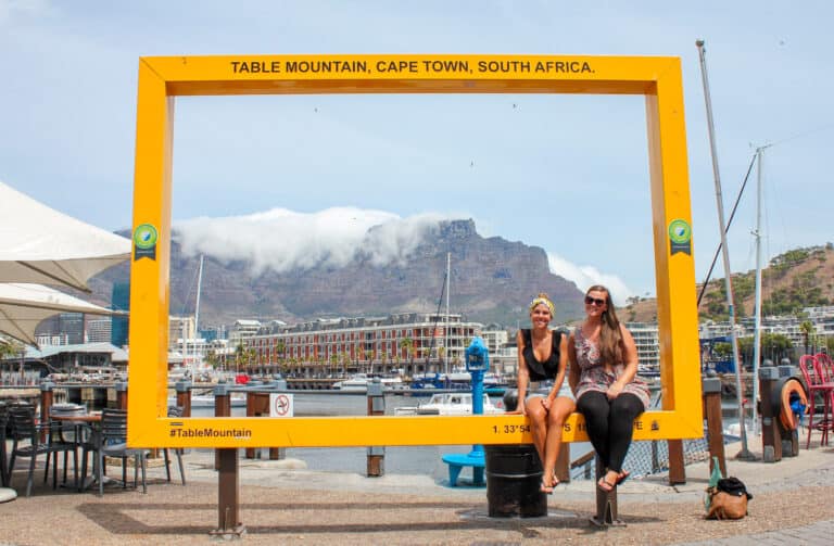 Two females sitting in a large yellow frame with Table Mountain in the background, during their travel to Cape Town