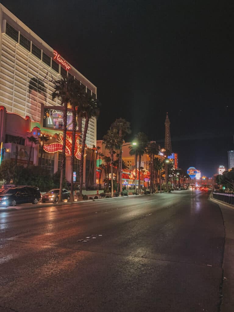 The Vegas strip at night. There isn't many cars on the road, palm trees line the middle of the road and all the buildings are lit up with bright neon lights
