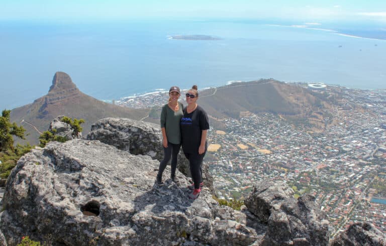 two females standing on large rocks facing the camera. Behind then you can see Cape Town and the Atlantic Ocean