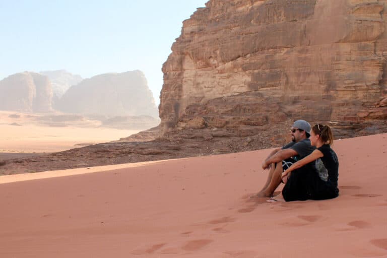 A man and women sit on the ground of Wadi Rum. The sand is a red color and they are looking into the distance