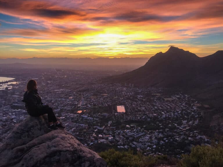 The sky is a bright pink, yellow and blue during a sunrise. I'm sitting to the left on a large rock, facing the view of Cape Town city.