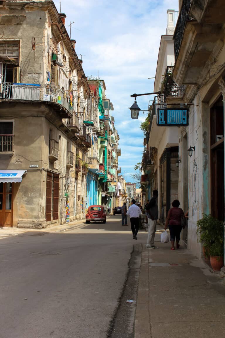 looking down a narrow street in Cuba, two old cars are parked on the side of the road and some of the buildings are painted bright blue and green