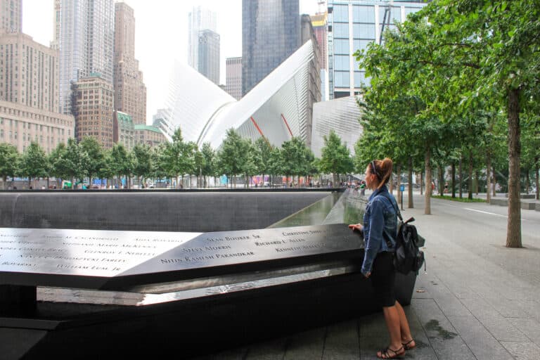 Elyse standing next to the black edge of the 9/11 memorial in New York City. Behind the memorial are skyscrapers and the memorial is lined with trees