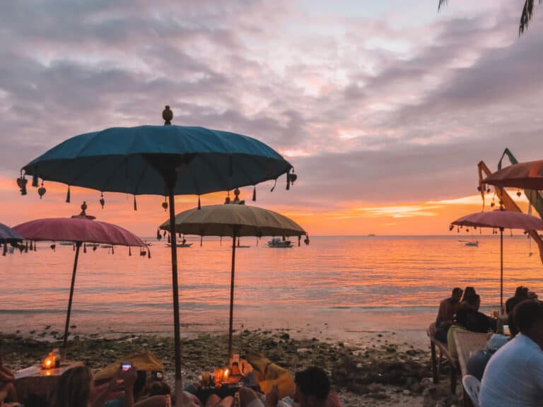 a restaurant on the beach with many customers sitting on bean bags under colorful umbrellas. The sun is setting in the distance making the sky very orange