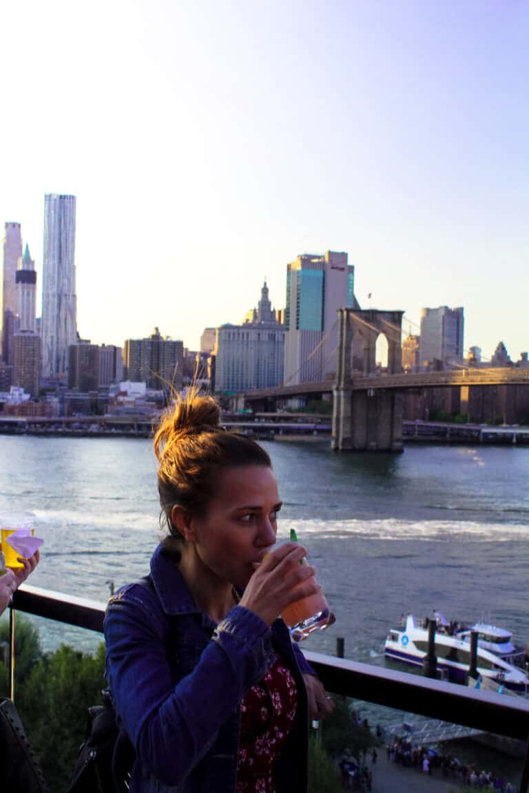 She's standing on a rooftop bar drinking a cocktail. In the background is Brooklyn Bridge and the city skyline. One of the experiences in New York City