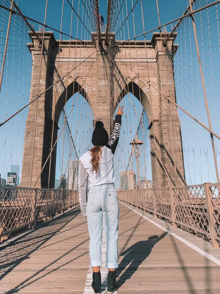 Elyse standing on the Brooklyn Bridge with her back to the camera. One arm is up in the air. A large amount of the bridge can be see in the distance.