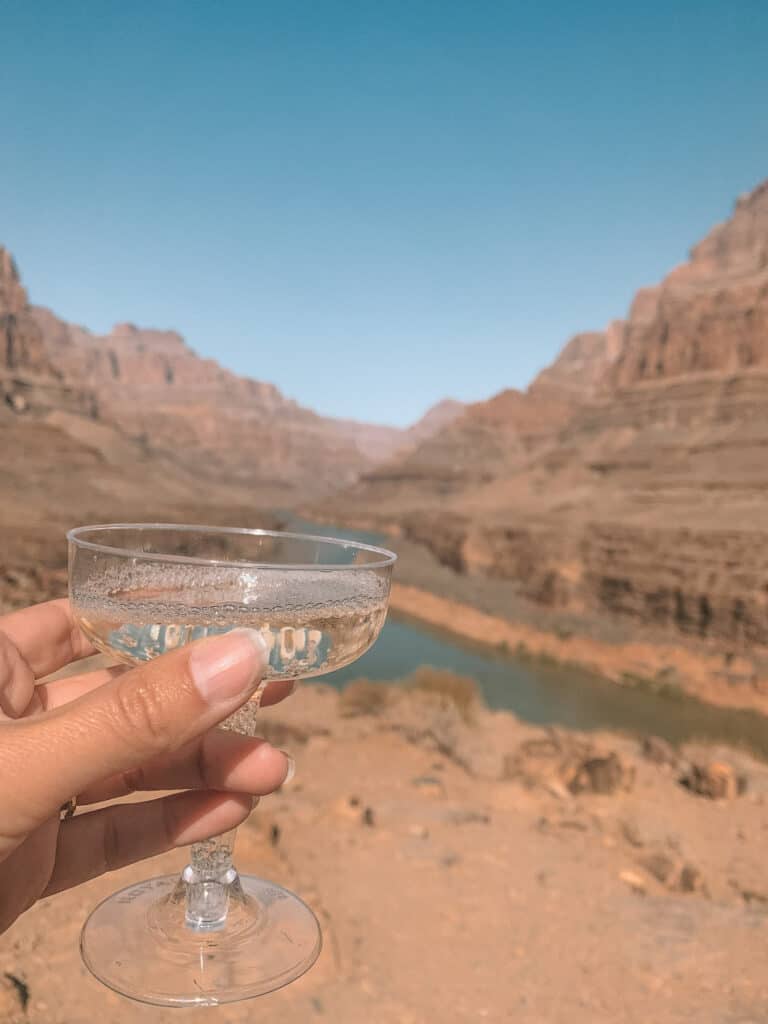 a hand holding up a champagne glass, in the background is the Grand canyon and view of the Colorado River