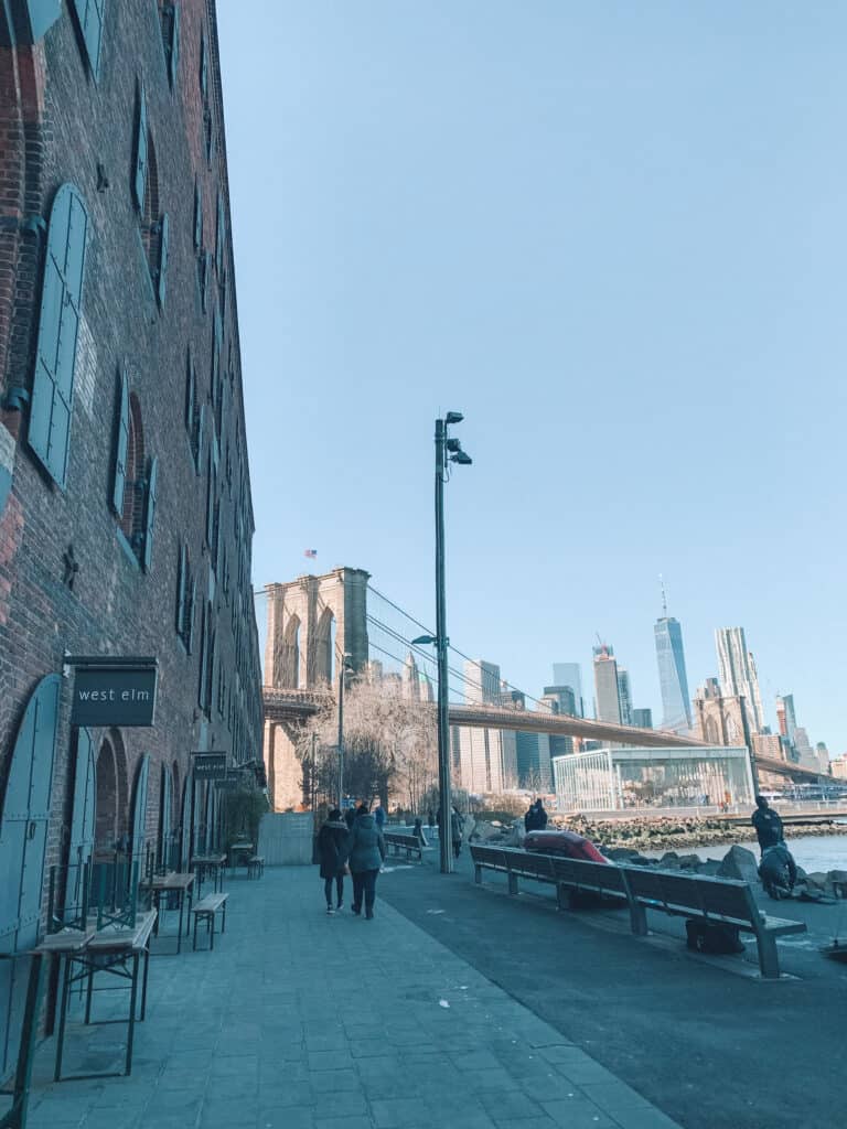 A quiet path of the edge of the river in Brooklyn. In the distant is a view of Brooklyn Bridge and the Manhattan skyscrapers