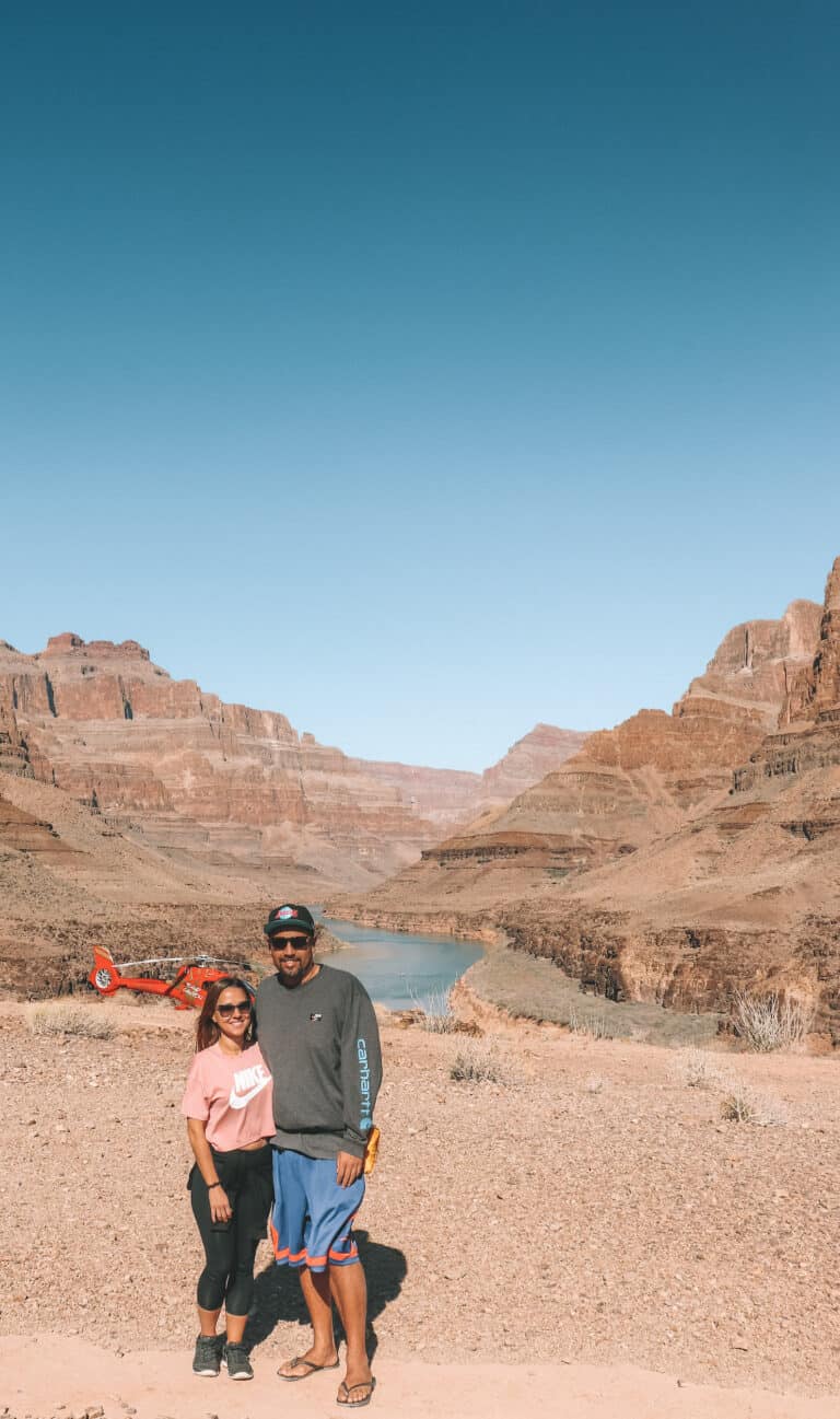 male and female standing inside the grand canyon with a red helicopter and the Colorado River in the background