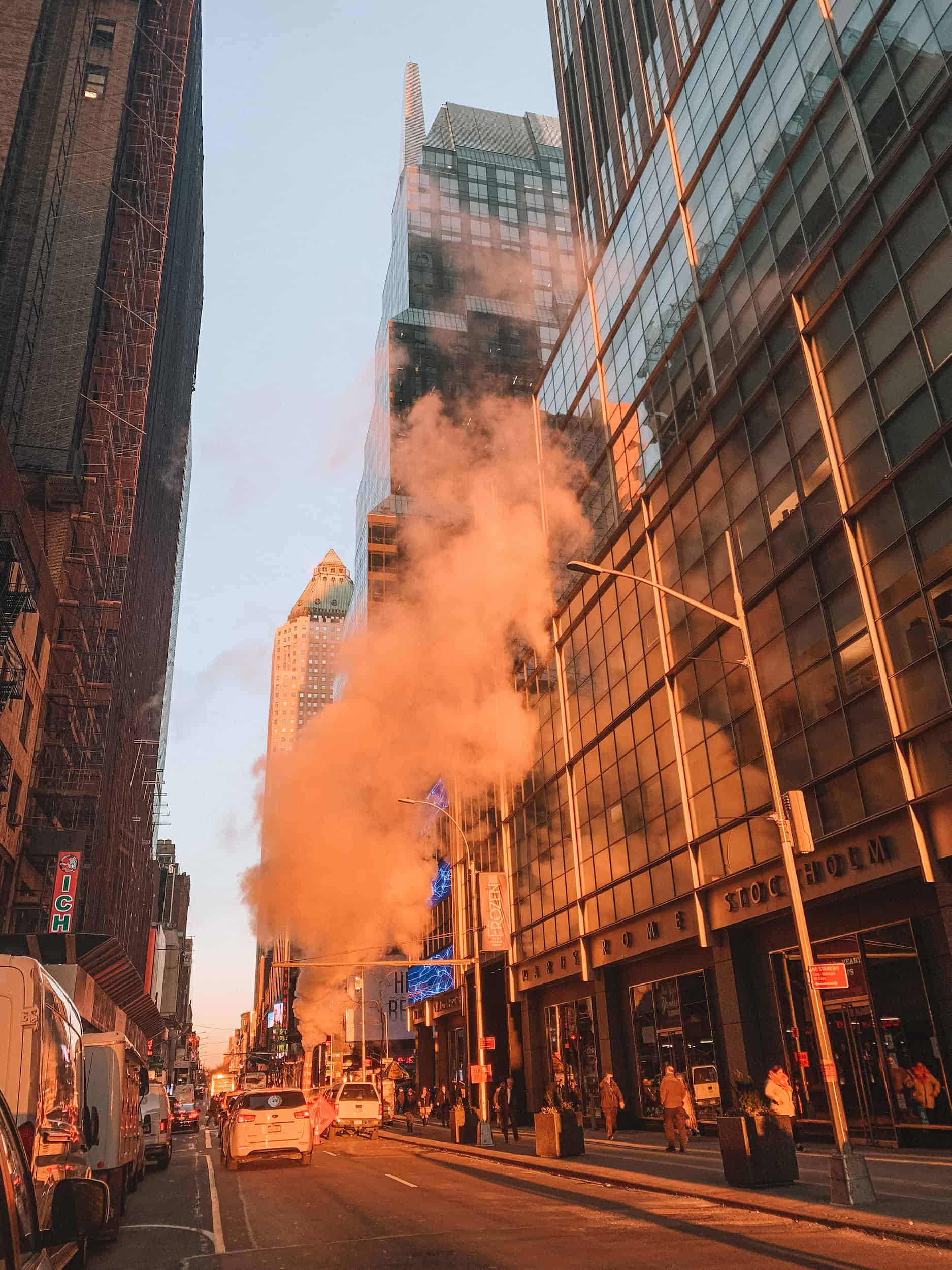 A two lane street in New York City glowing as the sun is coming up. A large amount of steam is coming up from the subway