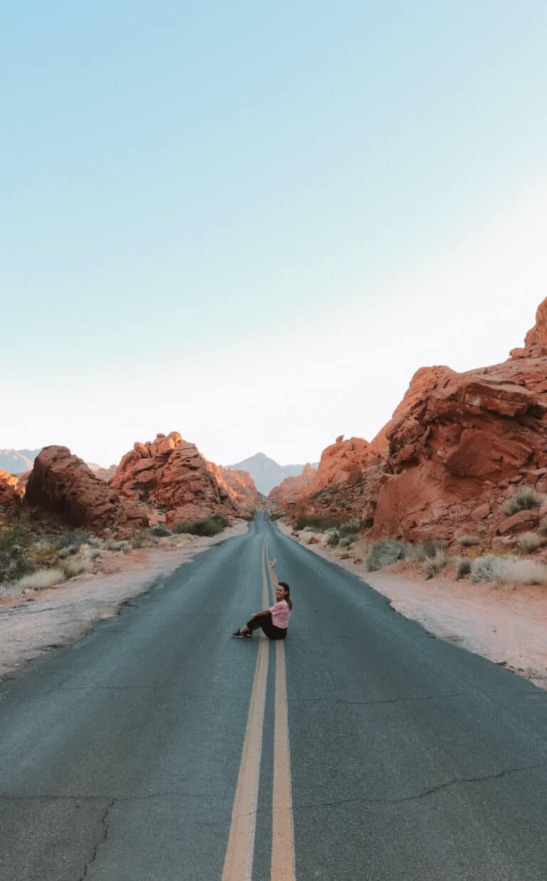 elyse sitting in the middle of a long and empty stretch of road during a Las Vegas day trip. red sandstone line each side of the road