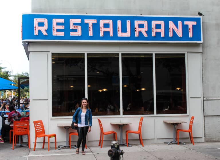 Elyse standing outside a cafe, orange chairs line the wall behind her and above is a neon sign saying restaurant, one of the experiences in New York City