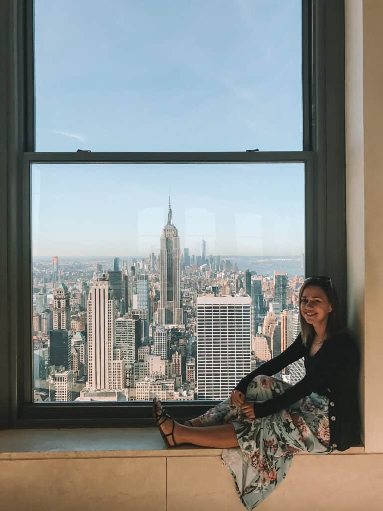 Elyse sitting on a windowsill, out the window is a view of new works buildings including the empire state building in the middle