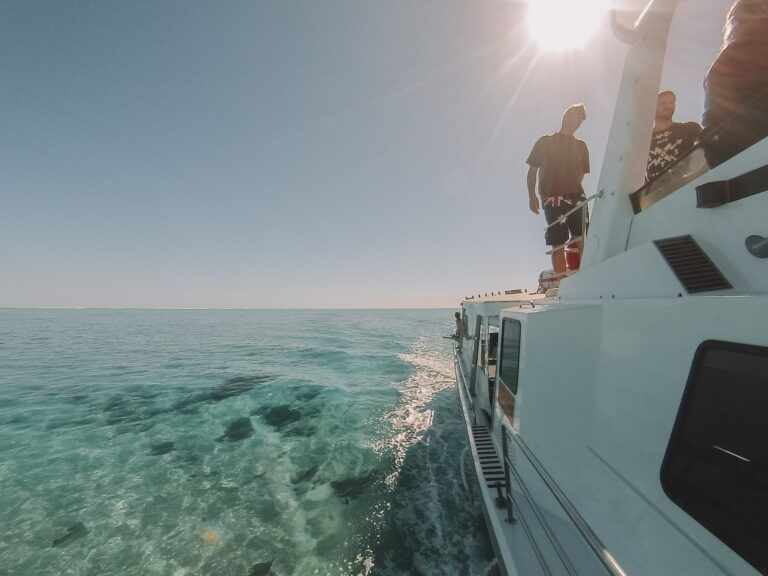 A boat out at sea on a sunny day, the image is shot looking down the side of the boat and there are a few people standing on the top deck waiting to swim with whale sharks.
