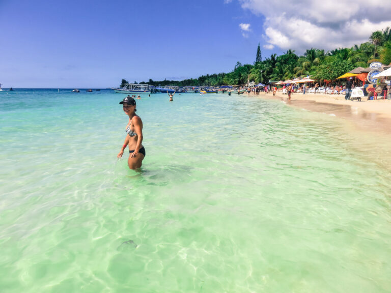 Elyse standing waste deep in the ocean on Roatan Island. The water is very clear and it’s a sunny day. The beach is lined with palm trees.