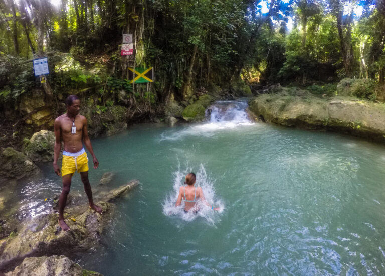 In the jungle a large swimming hole and Elyse is jumping into the water. A local Jamaican man is standing on the edge.