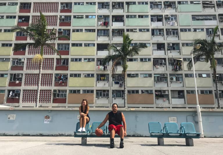 male and female sitting on the seats on the side of a basketball court. The male is bouncing the ball. In the back ground is a colorful apartment building and palm trees.
