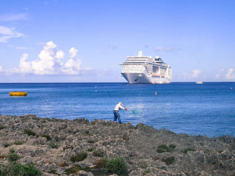A man fishing from the rocks on the waters edge in Grand Cayman, in the distance you can see a very large cruise ship.