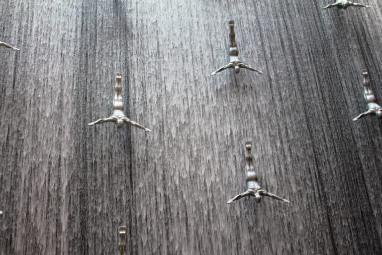 A display in the Dubai Mall of a water fall and silver metal statues. The statues are of men and it looks as if they are diving into the water.