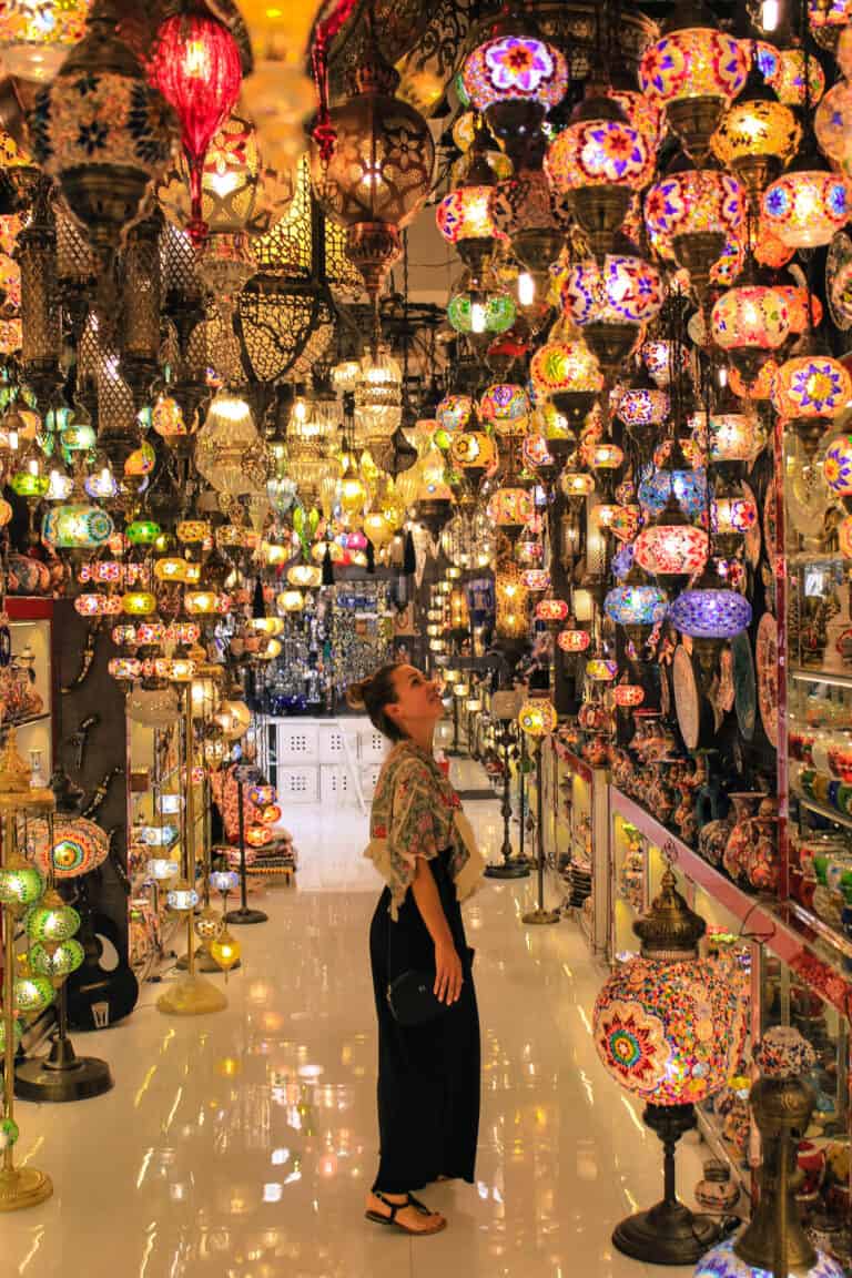 As this Dubai travel guide describes some of the shopping that is recommended in Dubai. This image shows Elyse standing in the middle of a store surrounded by bright and colorful lights. Some are lamps and some are hanging from the roof.
