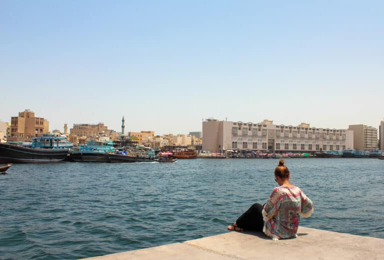 Elyse sitting on the edge of Dubais river, writing a travel guide. Across the water is many wooden boats and old buildings. It’s also a very sunny day.