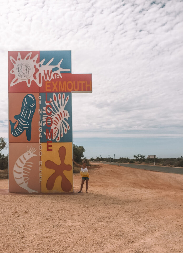 A large colorful sign on the side of the road, advising the beginning of Exmouth. Elyse is standing next to the sign with her arms out.