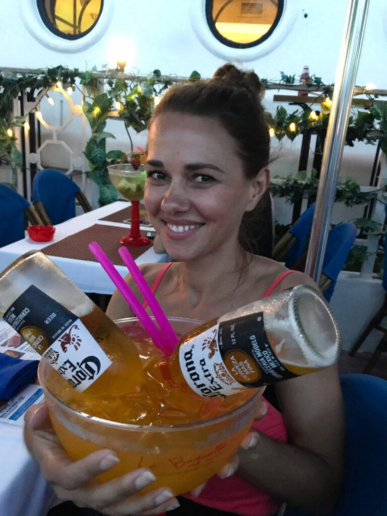 Elyse holding up a large cocktail with two Corona Beer poking out of the glass. She is smiling and the table behind her is empty.