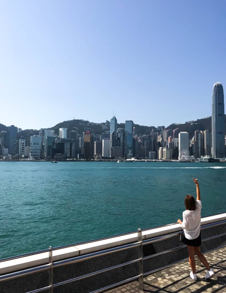 Elyse standing on the side of Hong Kong Harbor and across the water is the city skyline on a sunny day