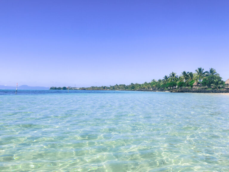 Taken when standing in the ocean, the image looks out to the clear blue ocean and in the distance is the shoreline in Samoa lined with palm trees.