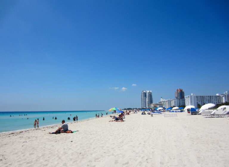 Looking down Miami Beach on a sunny day, there isn’t a cloud in the sky. The beach has people relaxing and the ocean is a bright blue color.