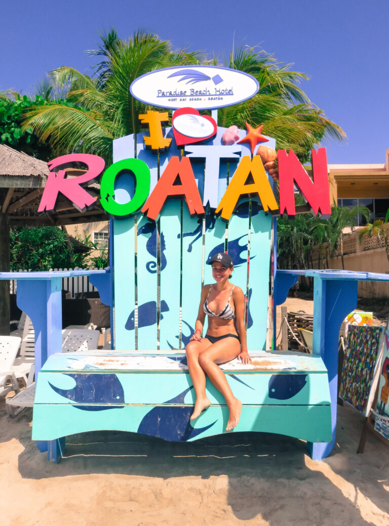 Elyse sitting on a large novelty seat, painting brightly with the island name Roatan on the top.