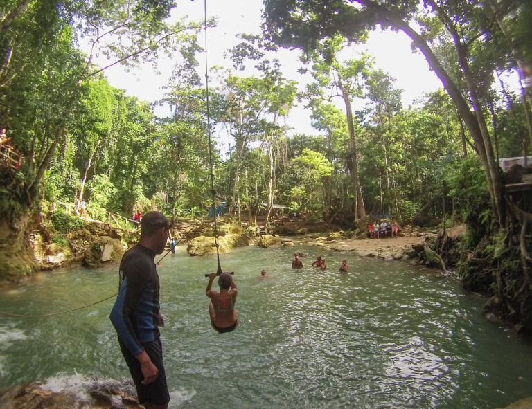 A large blue hole of water surrounded by the jungle. Elyse is holding onto a rope and she swings into the water.