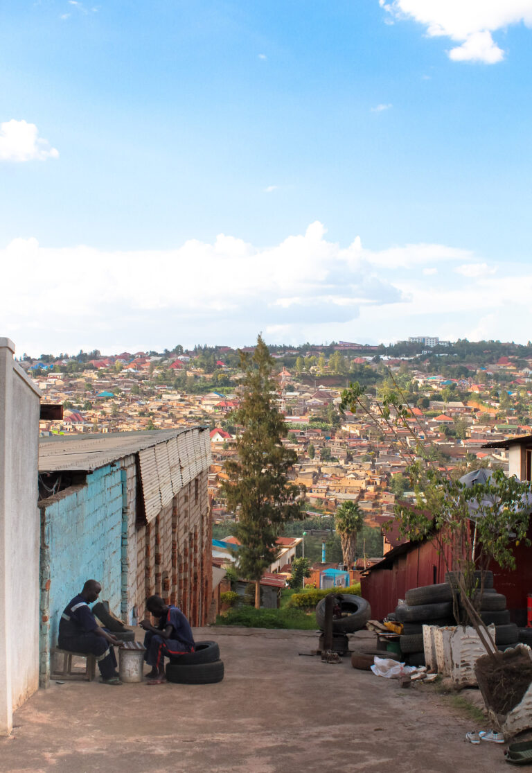 Looking down a side street in Kigali, the hills filled with buildings can be seen in the distance and two local men work on the side of the road.