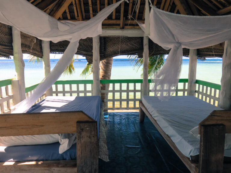 Two single beds are next to each other in a wooden Fale. In front of the Fale is a clear view of the beach.