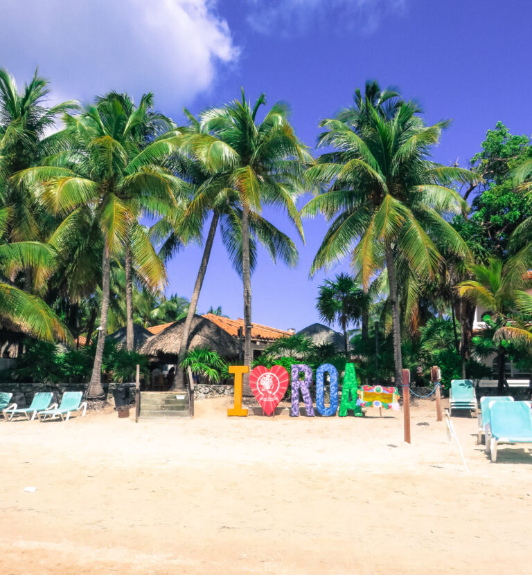 The sandy beach lined with palm trees and a large wooden sign of the letters I heart ROA.